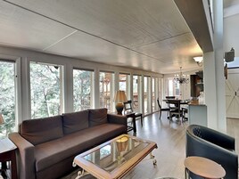 Un salón espacioso con un sofá marrón, una mesa de centro de vidrio y una mesa de comedor con sillas. Amplias ventanas ofrecen vistas a árboles fuera.