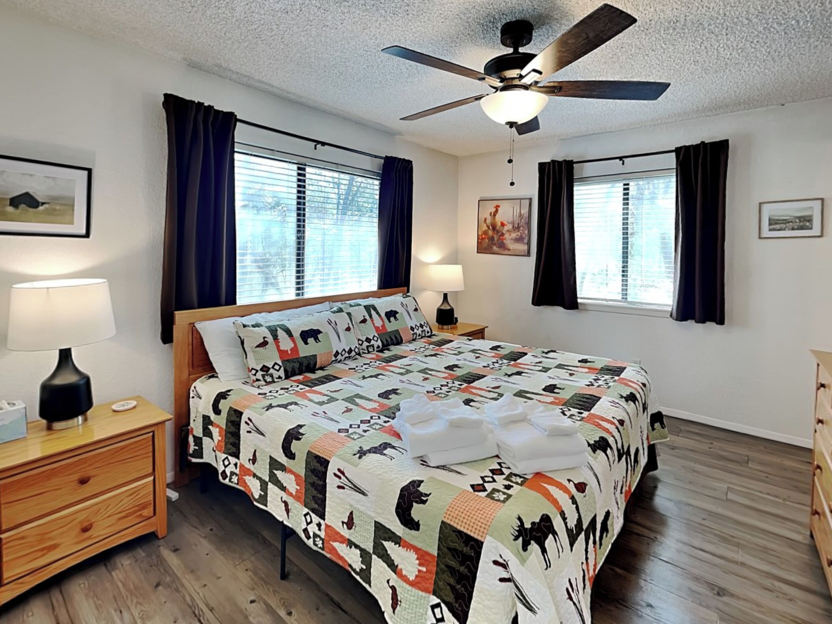 A bedroom with a bed, two bedside tables, a ceiling fan, and two windows with curtains.