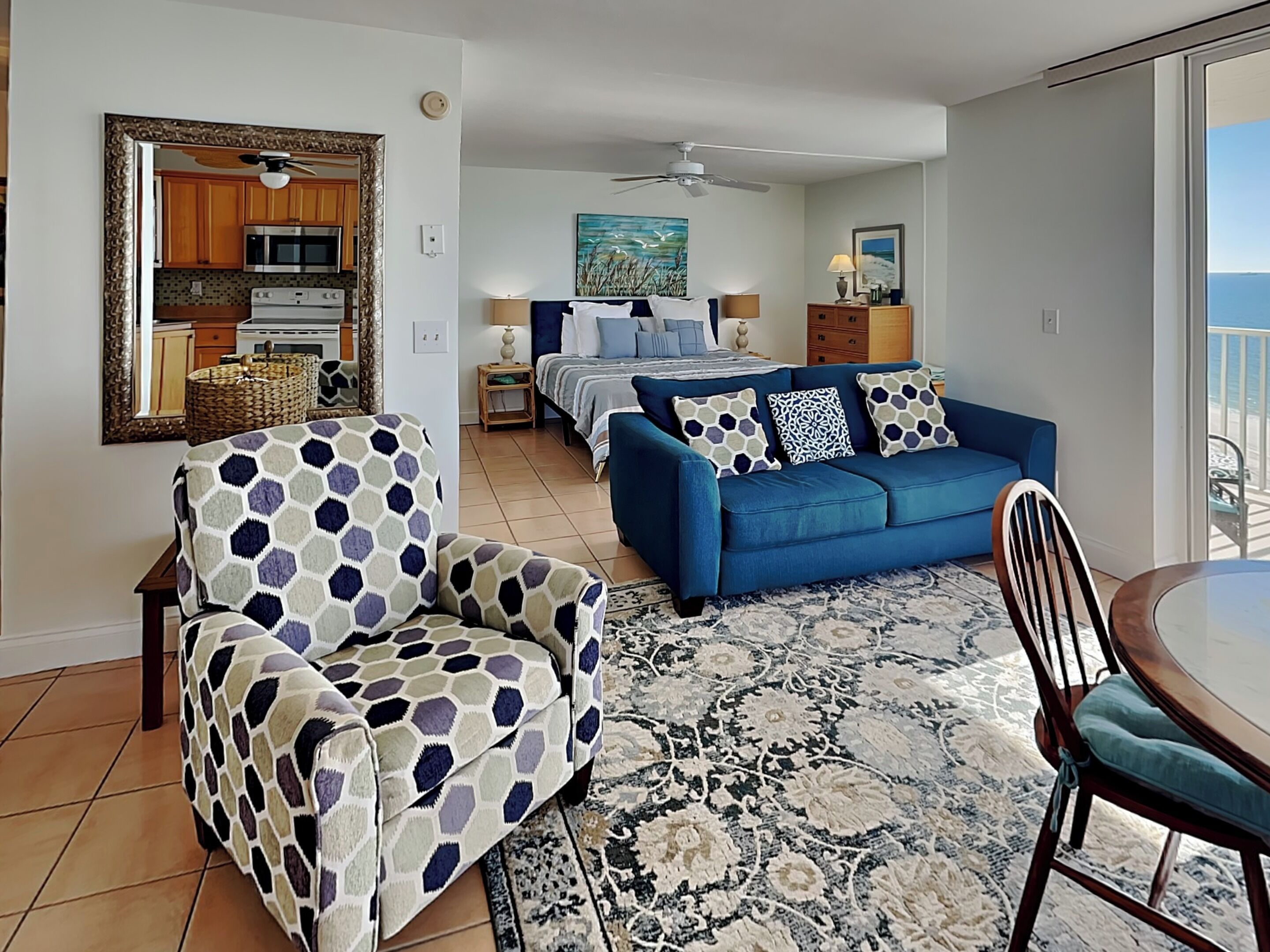 A living room with a sofa, armchair, and a dining table. There is a kitchen in the background with cabinets and a microwave.