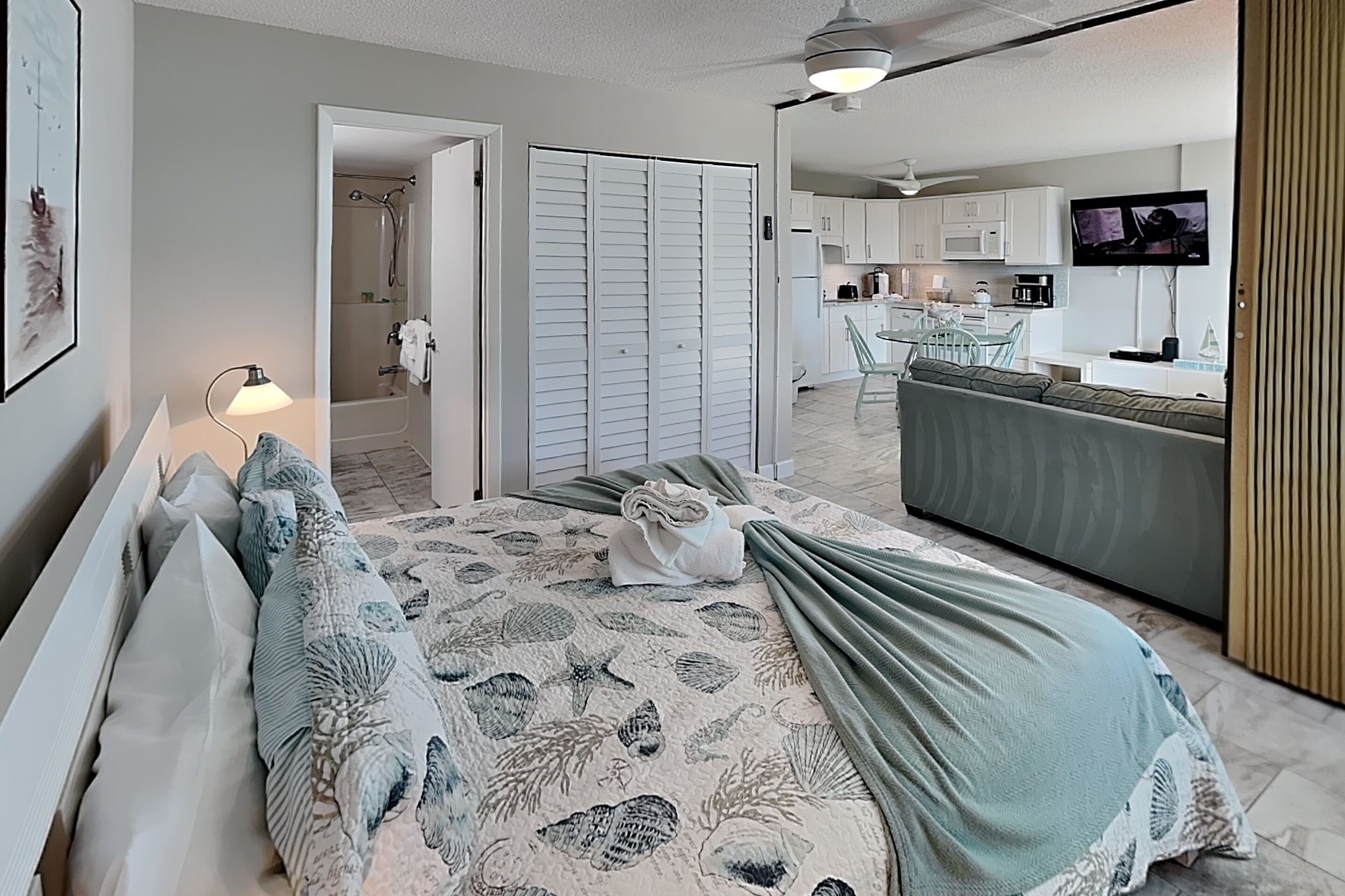 A bedroom with a bed, a nightstand, a lamp, a bathroom with a shower, and a kitchen area with a TV.