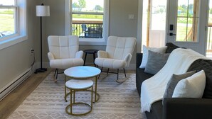 A living room with a sofa, two armchairs, a small round table, and a lamp. There are windows with views of a lawn and a building. - Bliss on the Bay (Birch Hill)