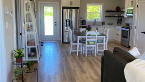 A spacious living area with a dark-colored sofa and a dining table with chairs. A kitchen area with a refrigerator, stove, and microwave is visible in the background. - Bliss on the Bay (Birch Hill)