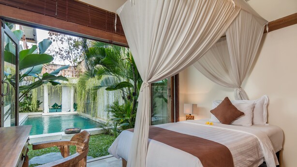 Ein Schlafzimmer mit Bett, Nachttisch, Stuhl und Blick auf einen Swimmingpool und Grünflächen.