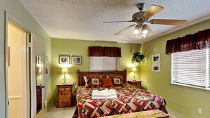 Ein Schlafzimmer mit einem Bett, Deckenventilator, zwei Nachttischen mit Lampen, einer Bank und einem Fenster mit Jalousien.