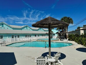 A pool area with a thatched umbrella, white Adirondack chairs, and a turquoise building with a white fence in the background. - Nemo Cay by Silver Sands Vacation Rentals (Corpus Christi)