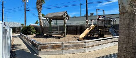 A playground with a slide and a wooden structure, surrounded by gravel and a wooden fence.