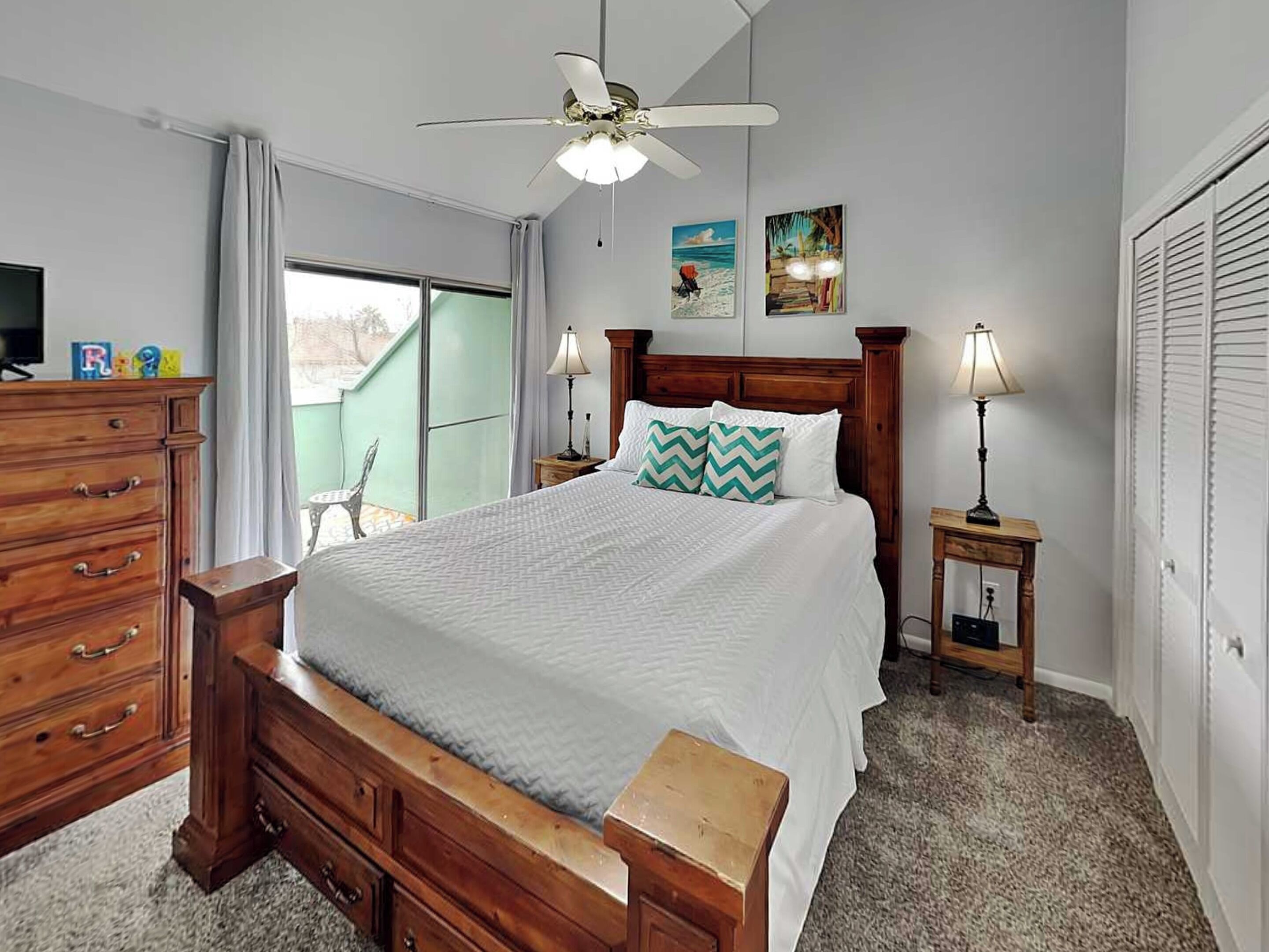 A bedroom with a bed, wooden dresser, ceiling fan, bedside lamps, and artwork on the wall.