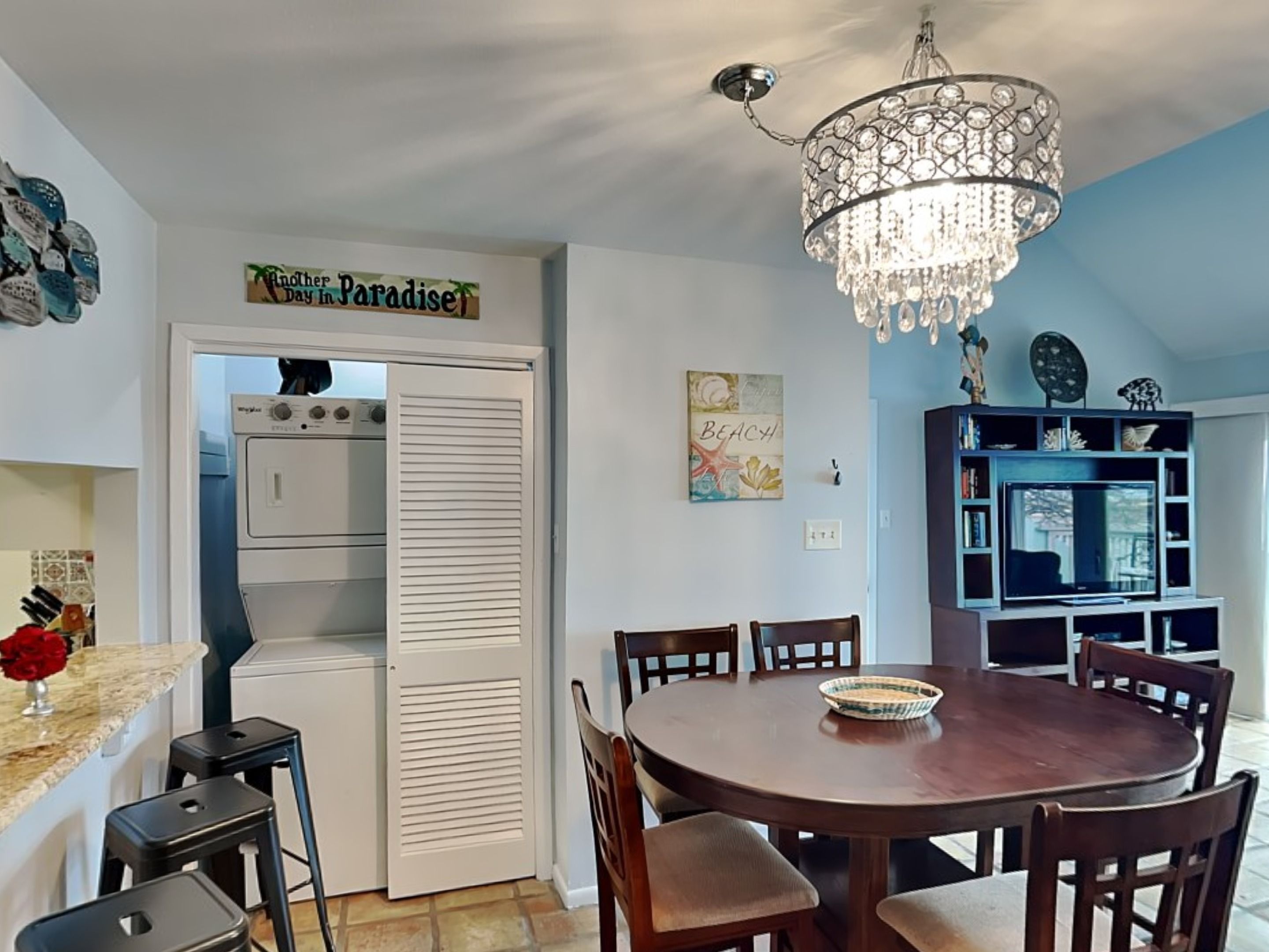 A dining area with a chandelier, a kitchen with a washing machine, and a sign that reads 'Another Day in Paradise.'