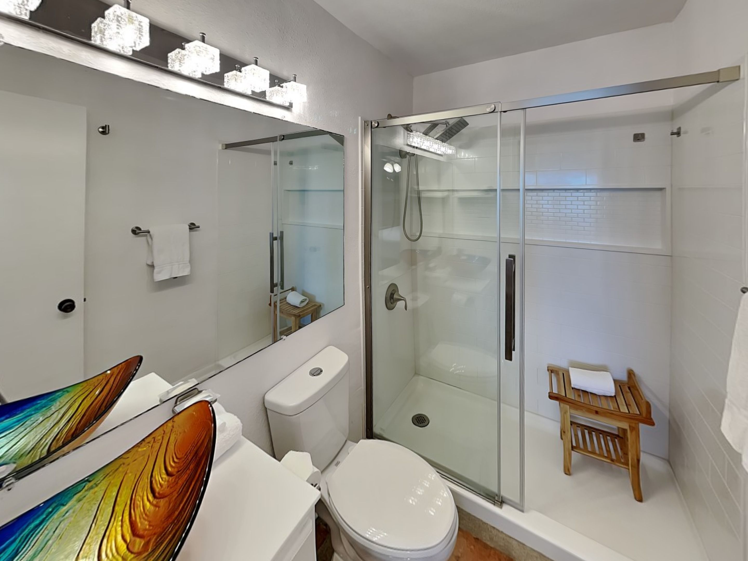 A modern bathroom with a glass-enclosed shower, a white toilet, a glass shower door, and a wooden chair.