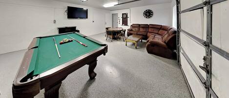 A pool table with balls, a sofa, a clock, and a television in a room with a garage door.