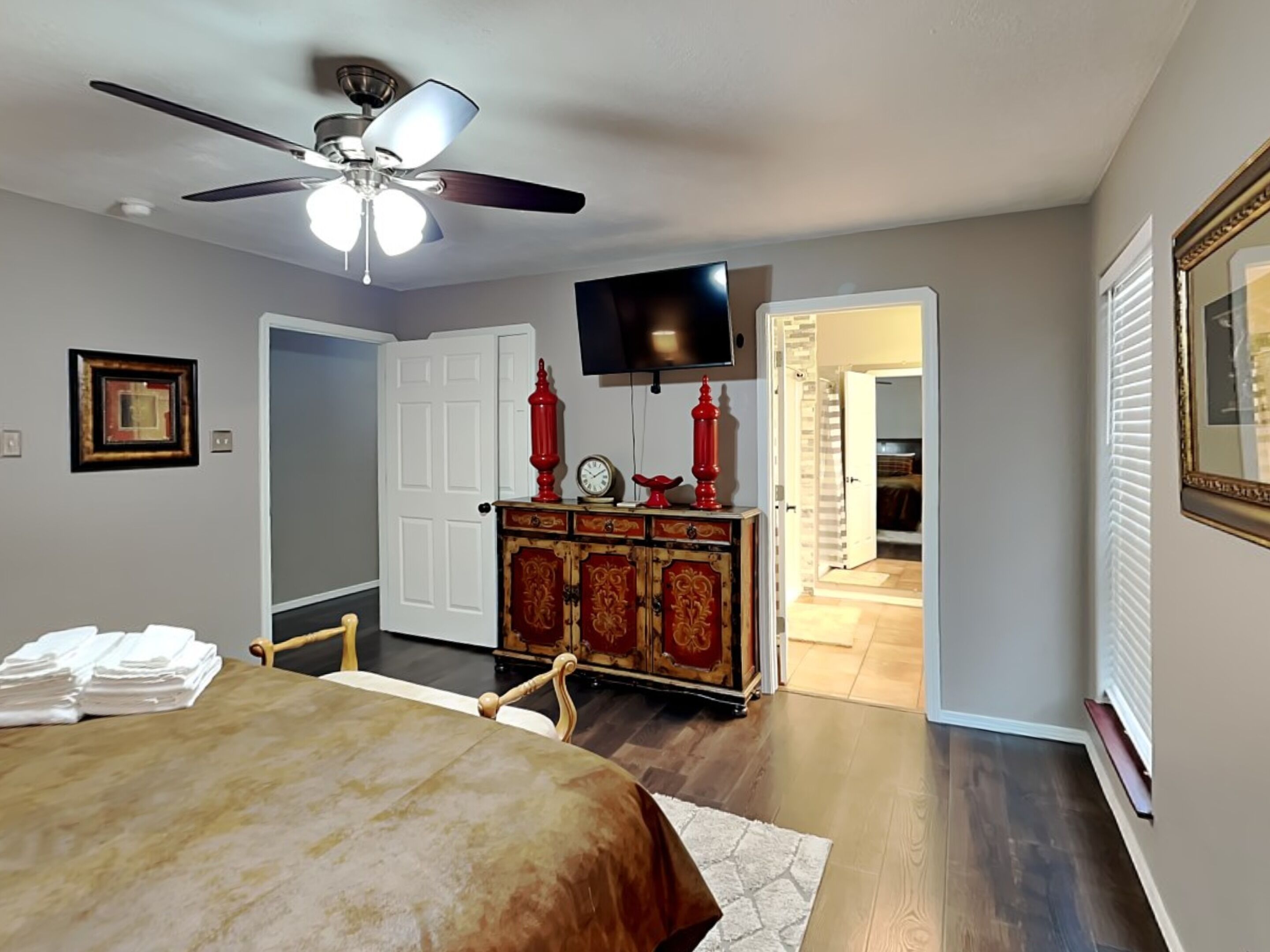 A bedroom with a bed, a ceiling fan, a dresser with red accents, a television, and a door leading to another room.