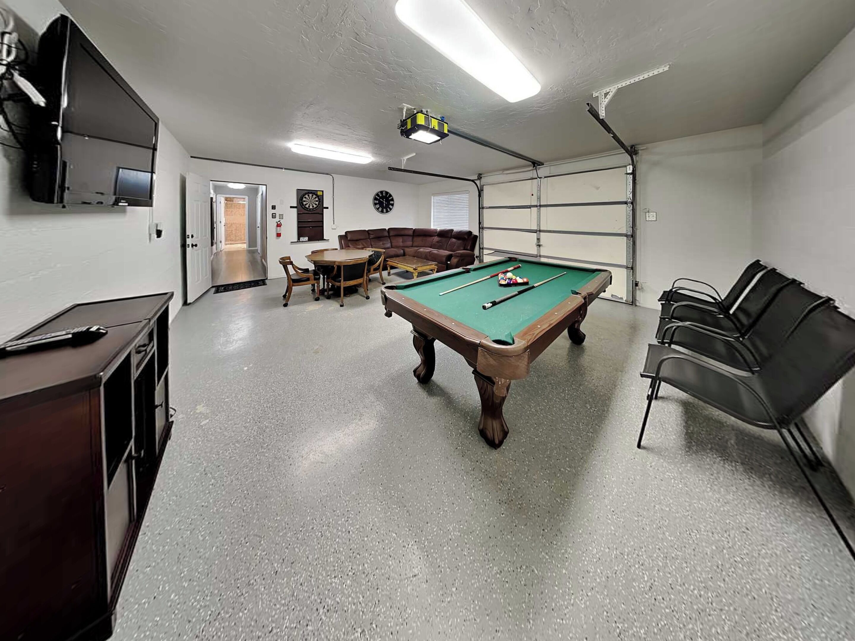 A pool table, chairs, a sofa, a television, and a clock in a room with a concrete floor and white walls.