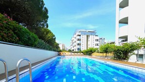 A swimming pool with a wooden deck, surrounded by greenery and modern buildings.