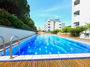 Une piscine dotée d’une terrasse en bois, entourée de verdure et de bâtiments modernes.
