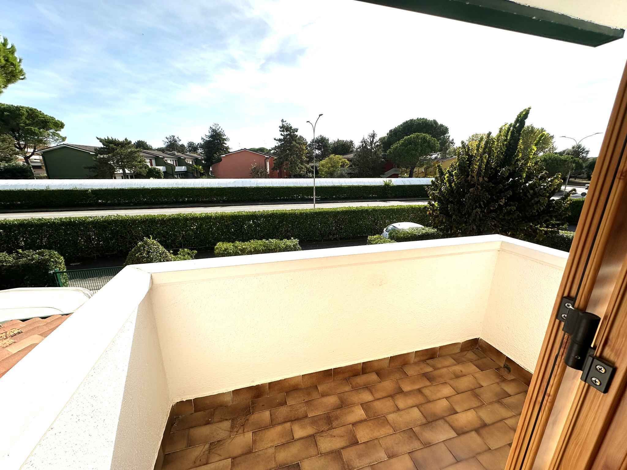 Un balcon doté d’un sol carrelé, d’une porte en bois et offrant une vue sur un arbuste taillé avec soin et sur des bâtiments en arrière-plan.