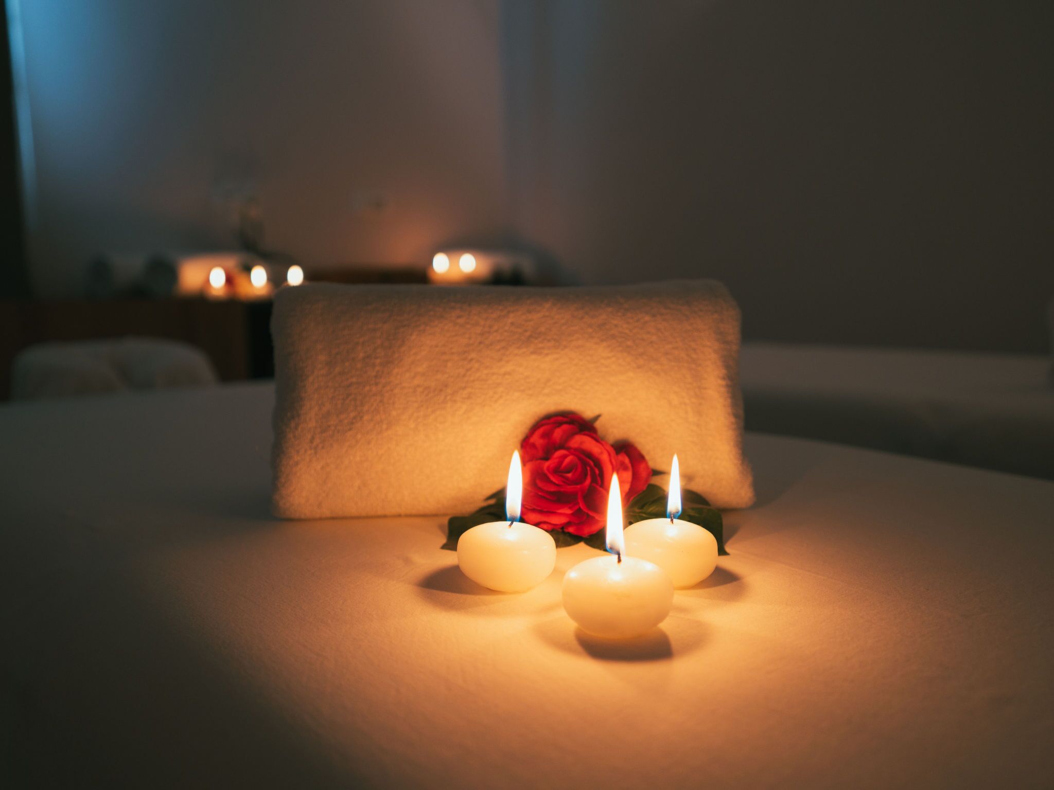 Ein Bett mit weißem Kopfkissen und drei brennenden Kerzen, eine rote Rose und eine Kerze auf einem Nachttisch.