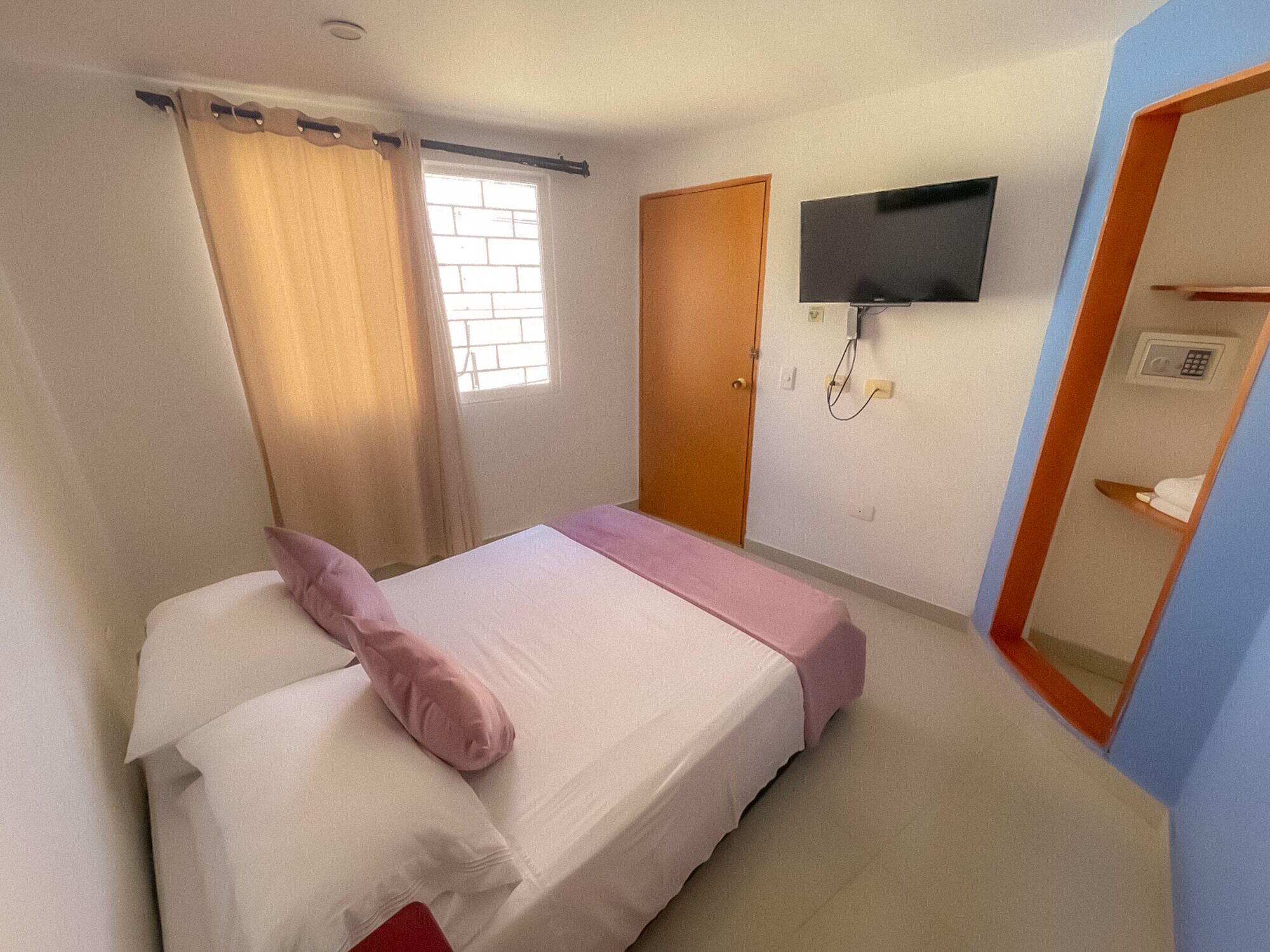 A bedroom with a bed, a TV mounted on the wall, a window with curtains, and a door.
