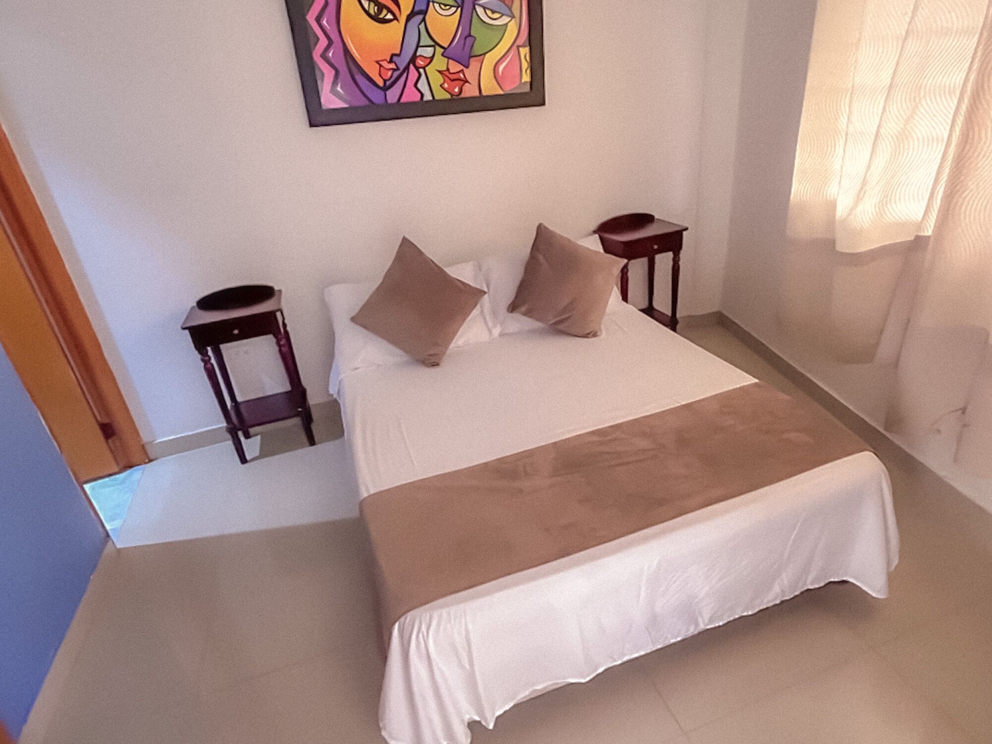A bedroom with a bed, two bedside tables, a painting on the wall, and a window with curtains.