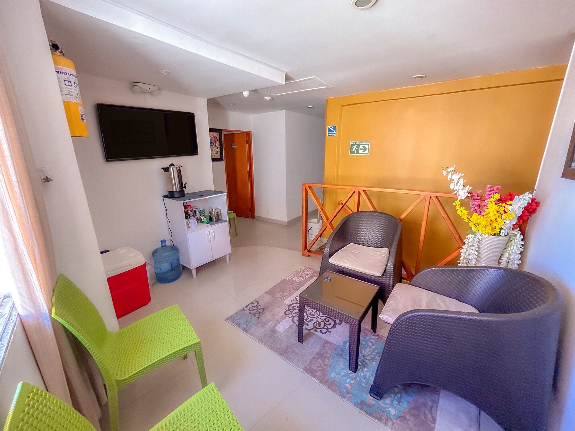 A compact living space with a flat-screen TV, a kitchenette, a wicker chair, and a small table.