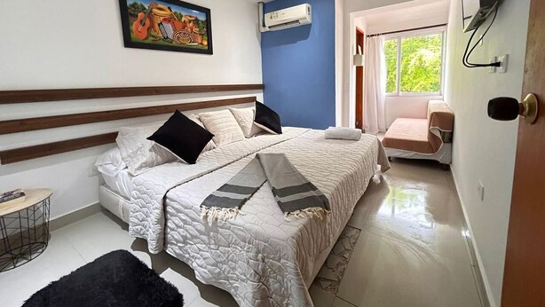 Un dormitorio con cama, una mesita, una silla, un equipo de aire acondicionado y un cuadro en la pared.