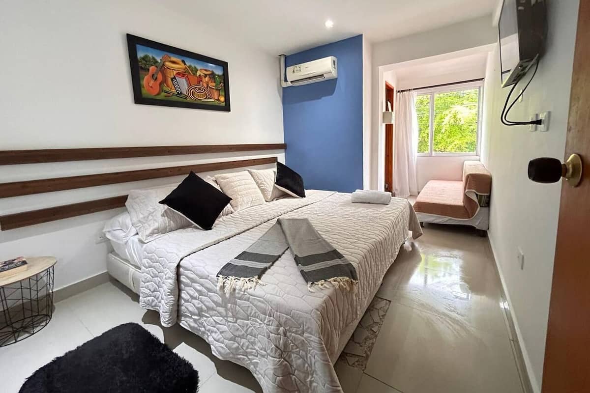 A bedroom with a bed, a small table, a chair, an air conditioning unit, and a painting on the wall.