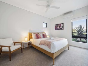 A bedroom with a bed, a chair, a bedside table, a ceiling fan, a window with a view, and a wall-mounted air conditioning unit.