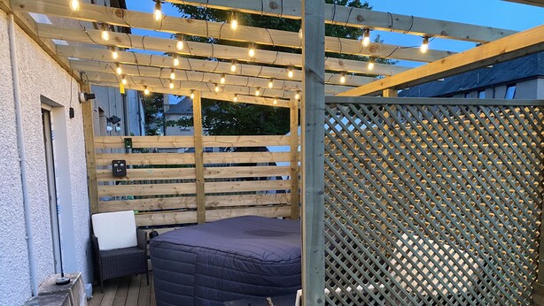 Un espace de détente extérieur accueillant, avec un banc rembourré et une petite table, installé sous une pergola en bois illuminée de guirlandes lumineuses.