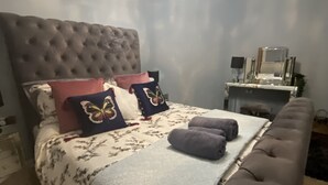 Une chambre à coucher avec une tête de lit capitonnée, un lit recouvert d’une literie à motifs floraux, des coussins décoratifs, une table de chevet avec une lampe et un miroir.