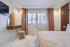 Quarto de hotel moderno com uma cama grande, televisão presa na parede, um banquinho pequeno e uma janela com cortinas.