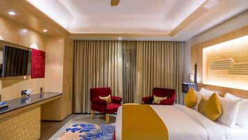 A hotel room with a bed, two red armchairs, a TV, a desk, and a large window with curtains.