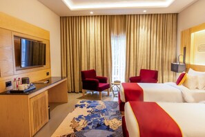 A hotel room with a TV, two red armchairs, a small table, a bed with white bedding, and a large window with curtains. - Ananta Spa & Resorts Jaipur (Jamwa Ramgarh)