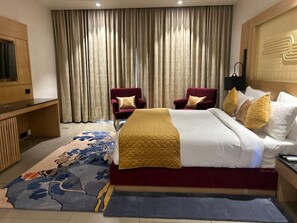 A hotel room with a large bed, two armchairs, a television, and a large window with curtains. - Ananta Spa & Resorts Jaipur (Jamwa Ramgarh)