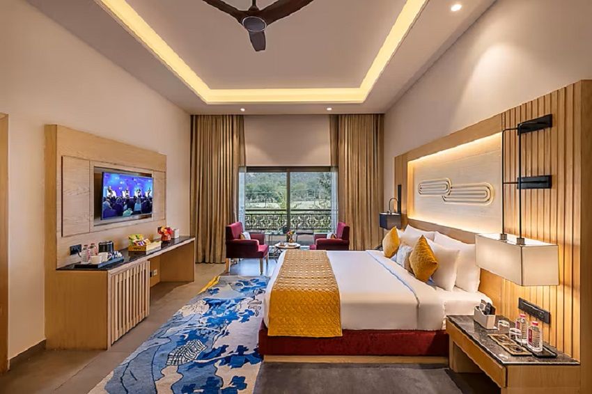 A hotel room with a bed, TV, desk, and a view of greenery outside.