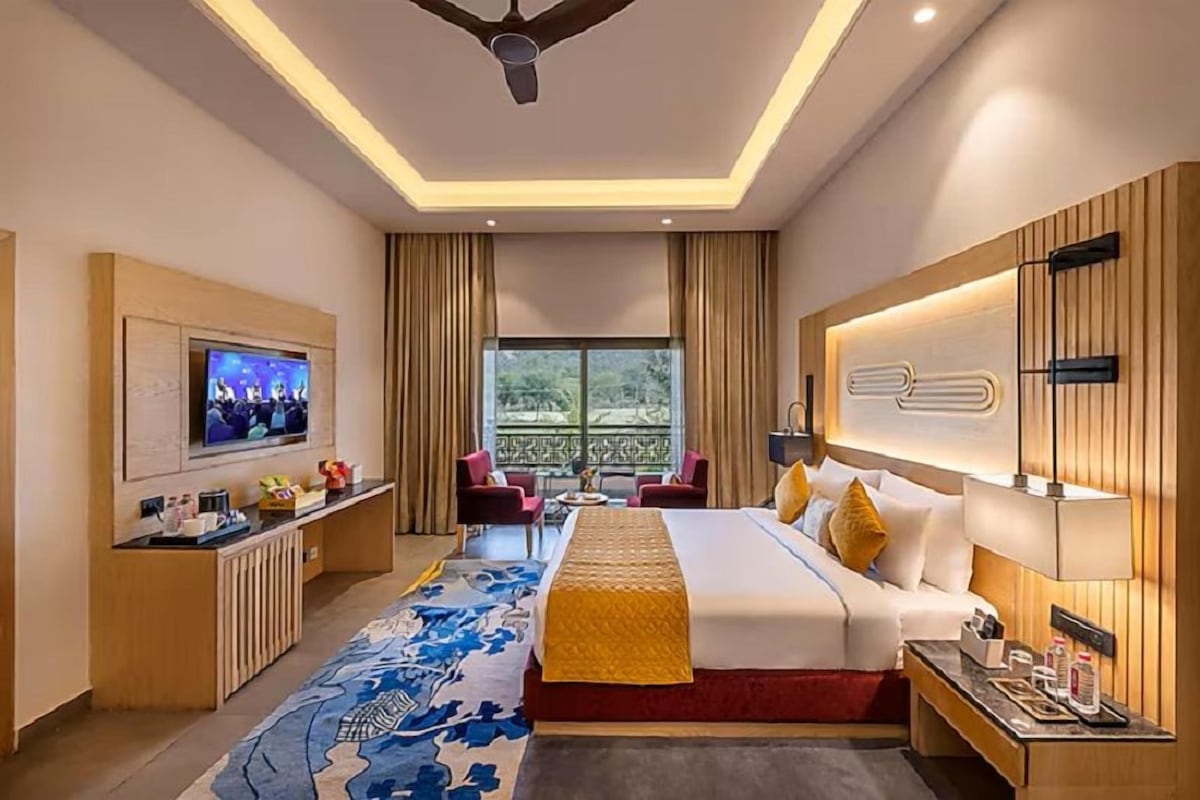 A hotel room with a bed, TV, desk, and a view of greenery outside.