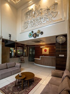 Um saguão de hotel com balcão de recepção, sofás, mesa de centro e um mural decorativo no teto.