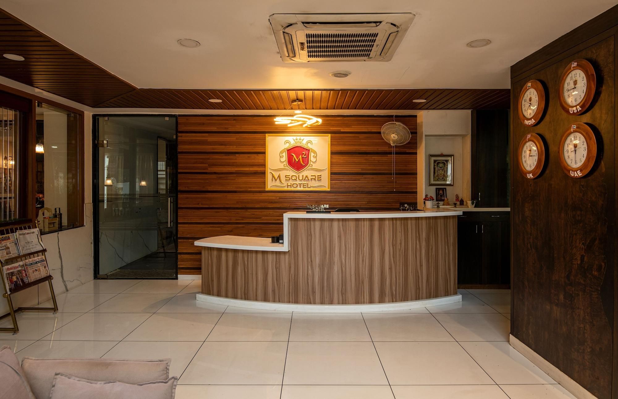 Un área de recepción con un mostrador de madera, un cartel del Hotel M Square y relojes que muestran la hora en varios países.