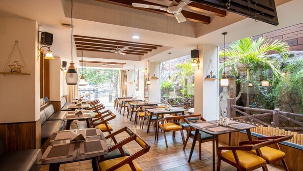 A restaurant with wooden tables and chairs, a high ceiling with exposed beams, and a view of greenery outside. - M Square Hotel-hitec City (Hyderabad)