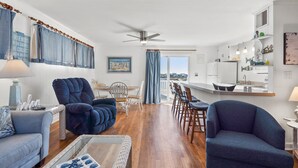 Un vaste espace de vie comprenant un grand fauteuil bleu, un canapé et une table à manger avec des chaises. On aperçoit également une cuisine avec un réfrigérateur blanc et des étagères au-dessus du plan de travail.