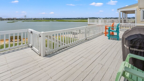 Une terrasse en bois avec une balustrade blanche donnant sur une étendue d’eau, un espace abrité avec une chaise, et une vue lointaine d’un château d’eau et de bâtiments.
