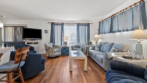 Un salon avec un plancher en bois, des meubles bleus et gris, un téléviseur et une fenêtre avec des rideaux.