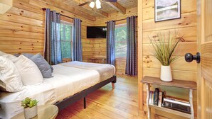 Een slaapkamer in een houten hutje, met een bed, plafondventilator, televisie en een boekenplank.