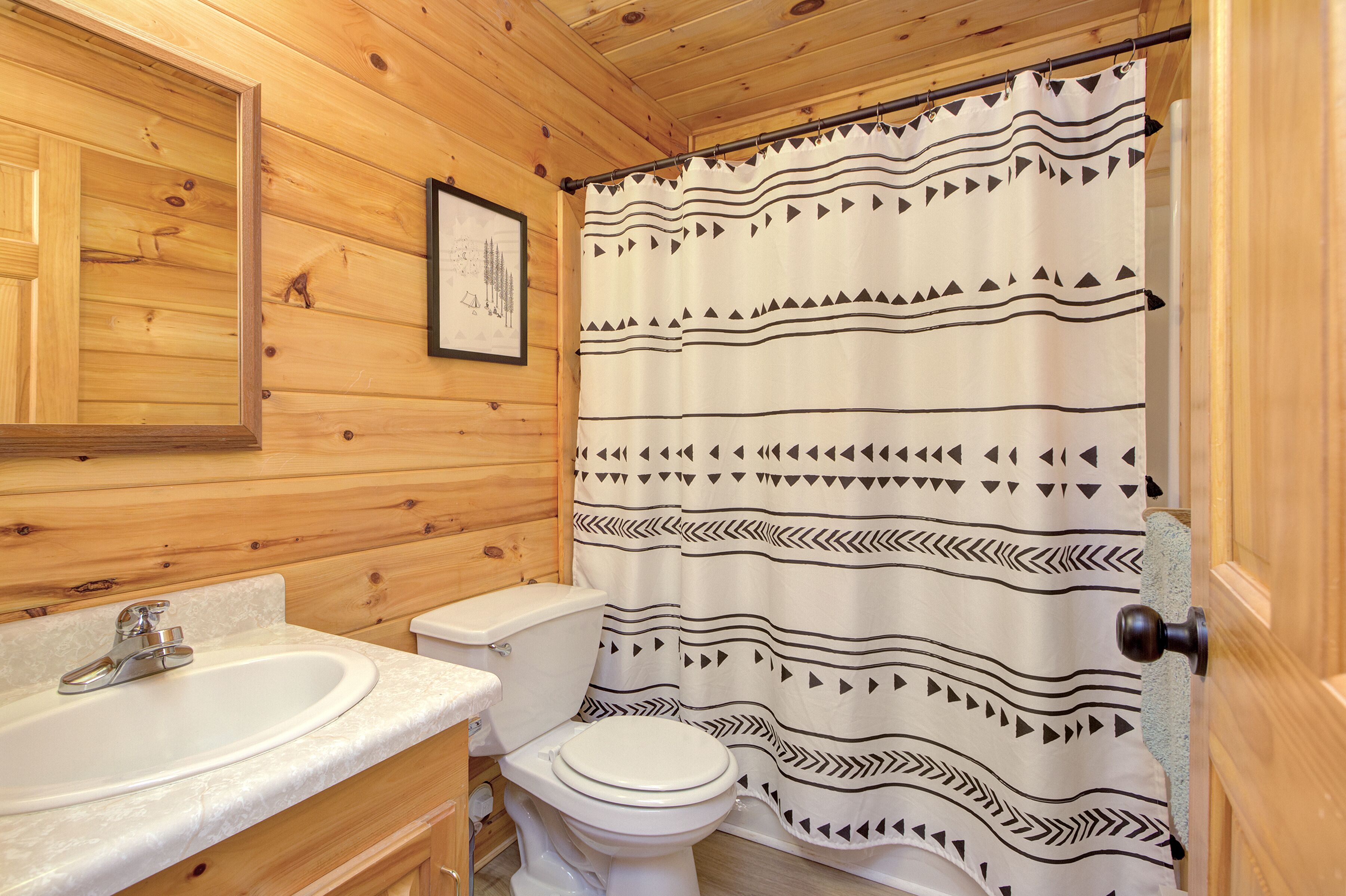 Baño con paneles de madera, un lavamanos blanco, inodoro y cortina de ducha con diseño geométrico.