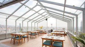 A rooftop restaurant with wooden tables and chairs, large windows, and a view of the city. - Hotel Bhrikuti Tara (Kathmandu)