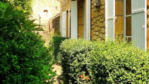 A stone building with a narrow pathway flanked by lush greenery and white shutters. - Beautiful home in Groléjac with WiFi (Groléjac)
