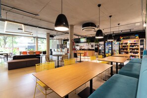 A modern cafe with wooden tables and yellow chairs, a blue sofa, and a kitchen area in the background. - Caza Noz by Viva - Apartamentos novos, com ótima estrutura e área comum próximo à USP e Butantã (São Paulo)