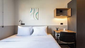 A hotel room with a bed, a desk, and a chair. There is a painting on the wall and a shelf with books.
