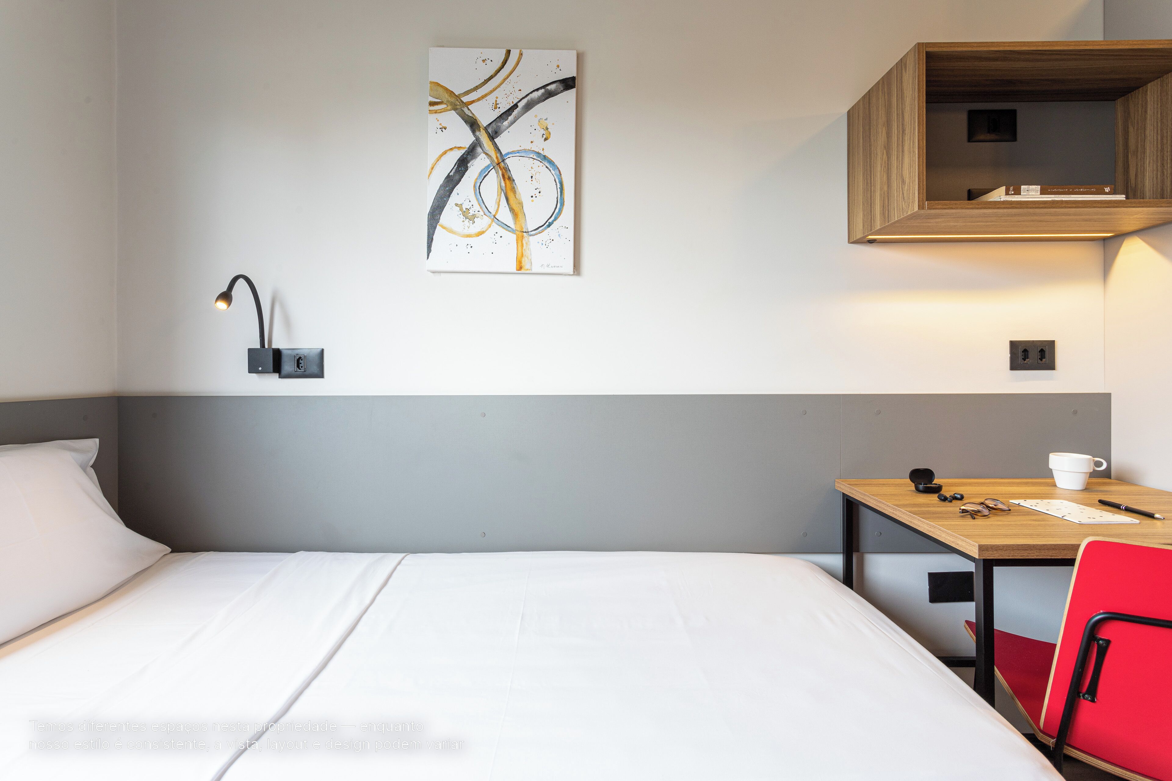 Habitación de hotel con cama, escritorio, silla, una obra de arte en la pared y estante con libros.