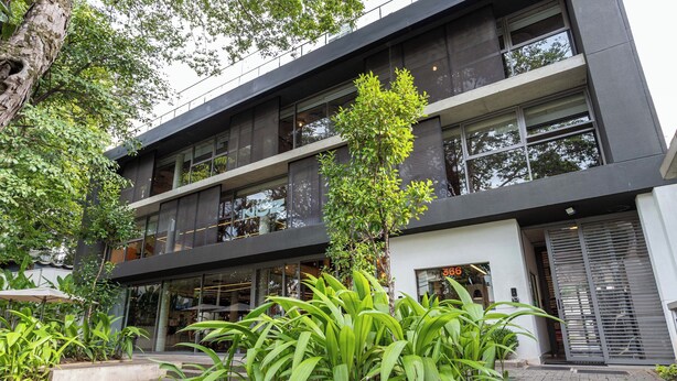 Un edificio moderno con fachada de vidrio, zona de estar al aire libre y abundante vegetación.