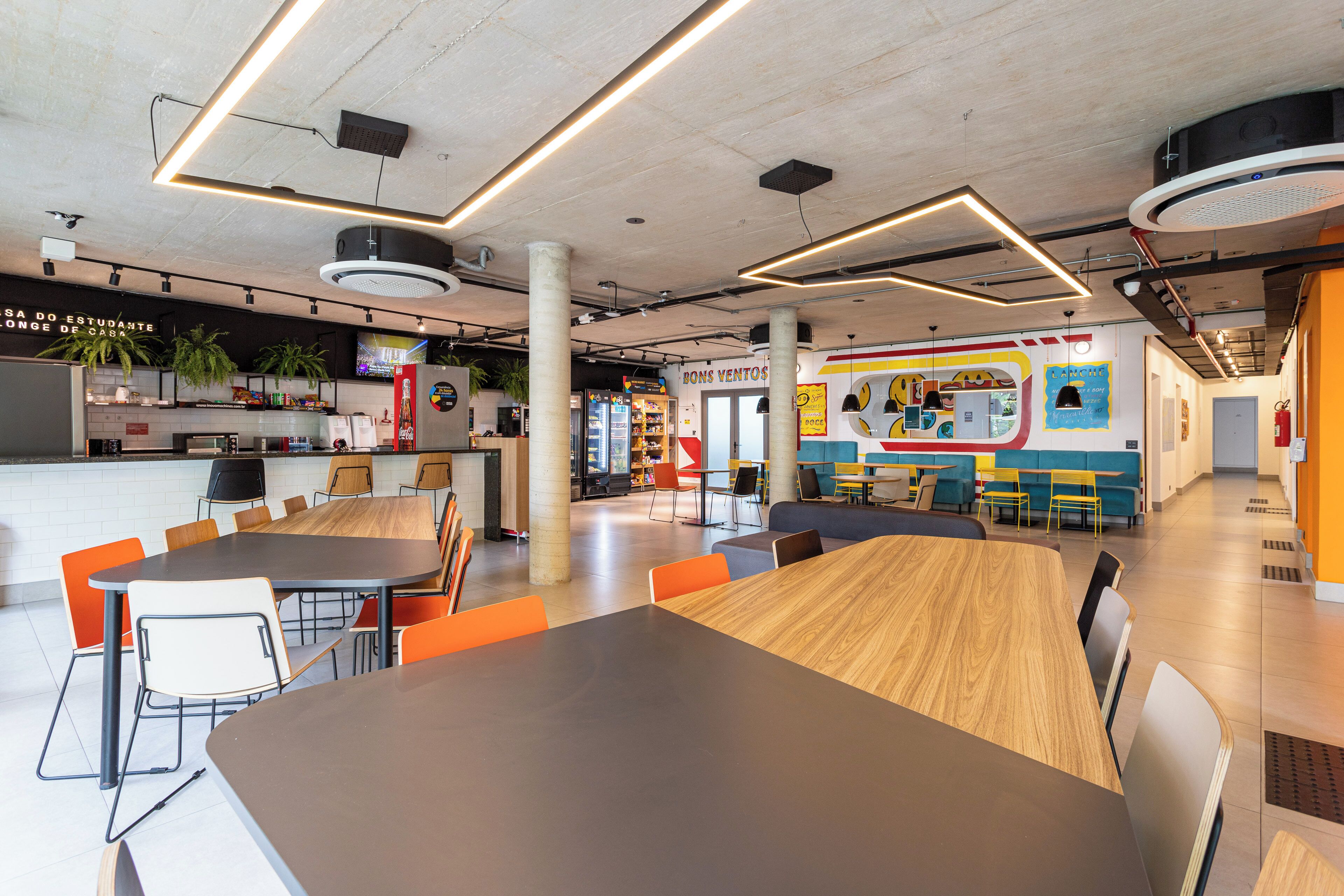 Una cafetería moderna con mesas de madera y sillas naranjas, un área de cocina y un mural colorido en la pared.