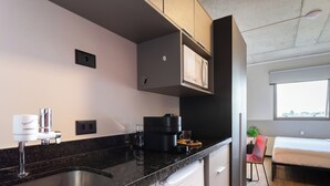 A modern kitchen with a built-in microwave, a coffee machine, and a sink. There is a small refrigerator and a window with a view of the city. - Caza Noz by Viva - Apartamentos novos, com ótima estrutura e área comum próximo à USP e Butantã (São Paulo)